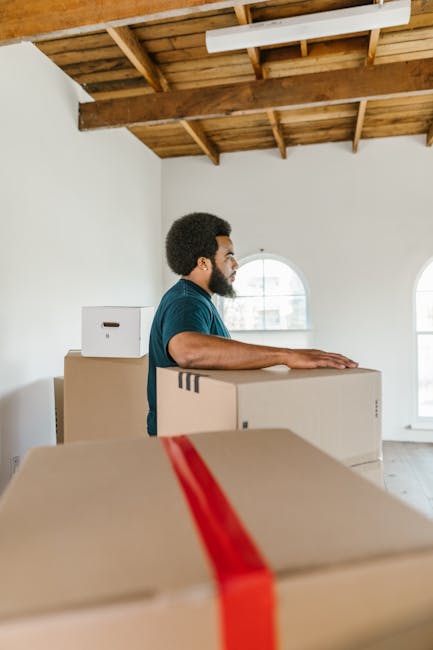 Two women are standing indoors surrounded by stacked cardboard moving boxes, some sealed with red packing tape. One woman, on the left, is holding a medium-sized box with a red and white label and wea