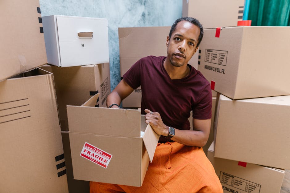 A man with a beard, wearing a black cap, white polo shirt, and jeans, is kneeling on the driveway next to an open cardboard box during a house removal process. He is using a black marker to label or r